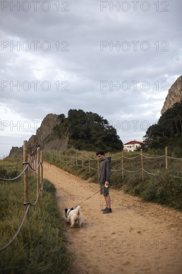 A serene scene of a man walking his dog along a sandy path in Ea, Basque Country, Spain The pathway is bordered by lush greenery and dramatic rock formations