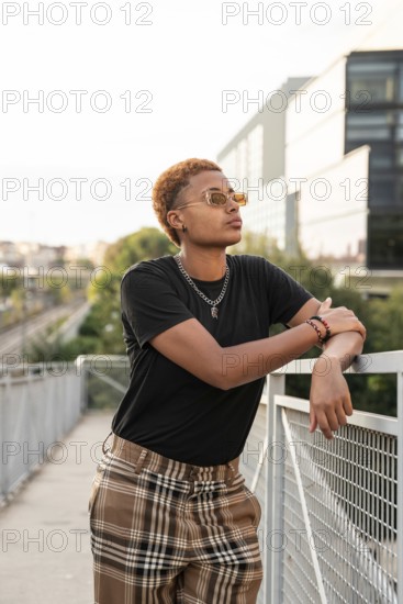 Young latin transgender with short hair and sunglasses, leaning casually against a railing in an outdoor urban setting, showcasing self-assurance and modern fashion