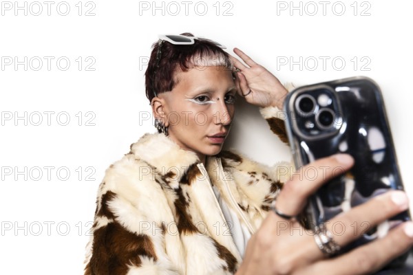 Stylish woman with bold eye makeup and piercings takes a selfie wearing a fur coat and sunglasses. The image captures a trendy, modern fashion statement and individuality