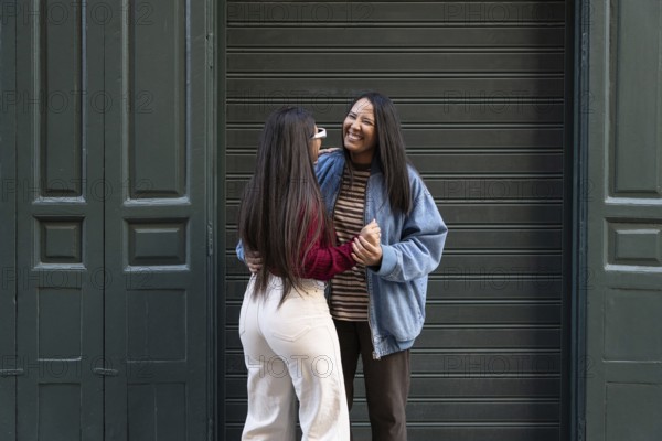 A joyful Latin lesbian couple stands in an embrace, laughing together in front of a green door. They exude happiness and connection in this candid outdoor moment