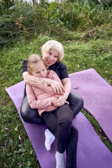 A joyful mother and daughter sit together on a yoga mat outdoors in a riverside. The lush, green surroundings and relaxed atmosphere highlight their affectionate bond and happiness