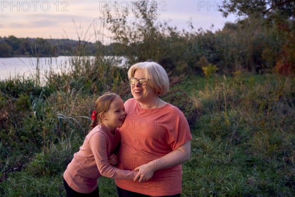 A joyful mother and daughter share a laugh in a serene outdoor setting by the riverside at sunset. They enjoy precious moments amidst lush greenery, creating lasting memories