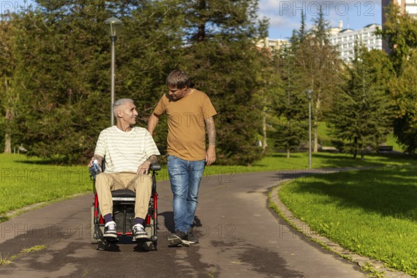 A young man walks alongside an elderly wheelchair user with cerebral palsy in a park, engaging in friendly conversation The path is surrounded by green trees and sunlight