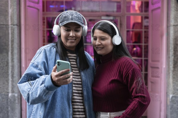 A latin lesbian couple listens to music with headphones while looking at a smartphone outdoors. They appear joyful and connected, sharing a moment on a casual day