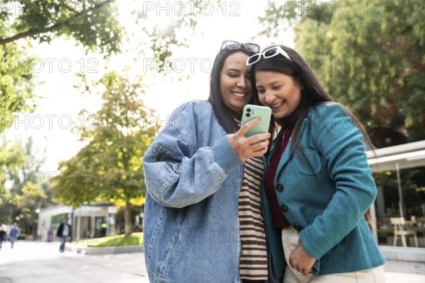 A joyous latin lesbian couple is seen sharing a heartwarming moment outdoors, smiling at a phone together. They are surrounded by lush greenery and sunlight