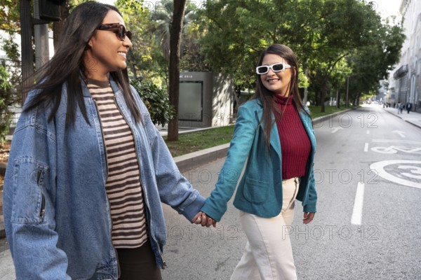 A joyful latin lesbian couple walks hand in hand down a sunlit city street, exuding happiness and love on a pleasant day, surrounded by lush greenery