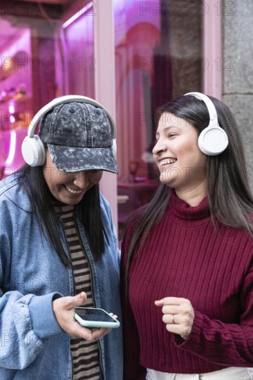 A latin lesbian couple enjoys music together with headphones. Smiling and relaxed, they share a joyful moment outdoors with vibrant colors around them, reflecting happiness and connection