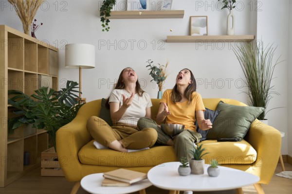 A joyful lesbian couple sits on a vibrant yellow sofa, tossing popcorn in the air and laughing together The modern, cozy living room radiates warmth, love, and happiness