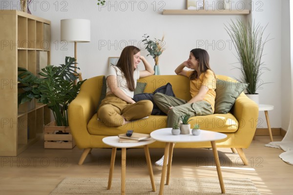 A cheerful lesbian couple enjoys a cozy moment on a yellow sofa The room is filled with greenery and soft lighting, highlighting the warmth of their connection