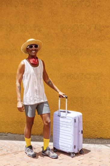 LGBTQIA+ man stands joyfully beside a suitcase, dressed vibrantly and ready for a holiday. A celebration of diversity and travel. Bright orange background enhances the lively mood