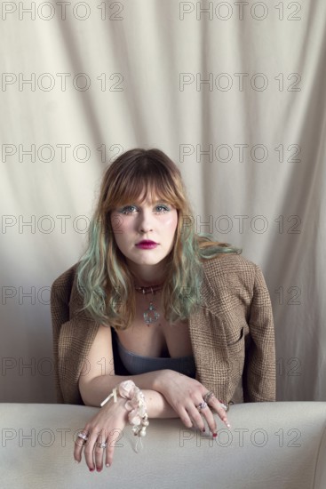 A woman with colorful hair leans on a sofa, wearing a brown blazer and eye-catching jewelry The neutral background accentuates her fashionable, trendy look