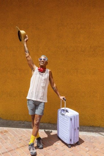 LGBTQIA+ man joyfully preparing for a holiday, standing with a suitcase against a vibrant orange wall. Their expression exudes excitement and pride