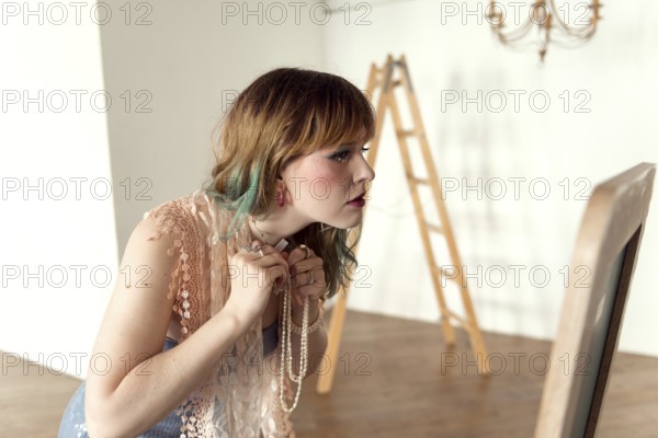 A woman in a lace top examines her reflection while wearing layered jewelry The minimalist room features a wooden ladder and chandelier, adding an artistic touch