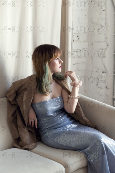 A stylish woman in a shimmering blue gown and brown coat sits thoughtfully on a beige couch Natural light highlights her outfit and creates a serene atmosphere