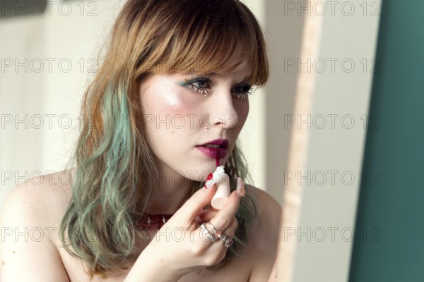 A woman carefully applies bright pink lipstick in a well-lit room Her wavy hair is styled casually, and she wears minimal jewelry The natural light enhances the vibrant makeup