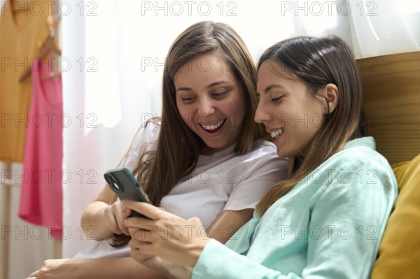 A joyful lesbian couple relaxes together while looking at a smartphone, sharing laughter and happiness This image reflects authentic love and togetherness in an LGBTQIA+ relationship