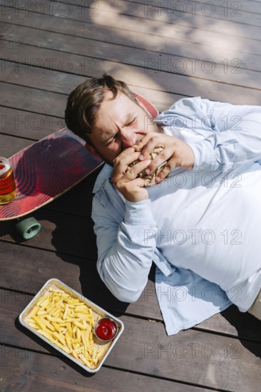 A person enjoys a barbecue meal outdoors, lying beside a skateboard. The scene includes fries with ketchup and a refreshing drink, capturing a laid-back, casual vibe