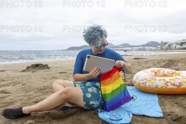 A transgender man with blue hair sitting on a beach, putting a tablet inside a rainbow pride bag. He's dressed in a blue shirt and floral shorts, symbolizing a tranquil moment by the sea