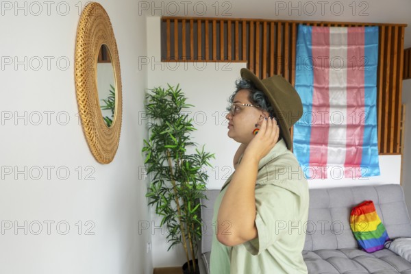 A young transgender person wearing a hat looking into a mirror reflection, stands near a couch in a room decorated with a transgender pride flag