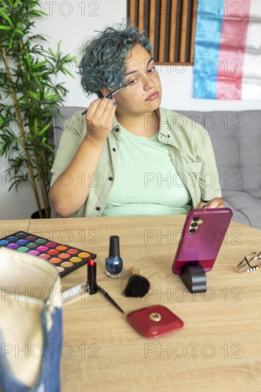 A transgender man meticulously applies makeup while recording a tutorial with his mobile phone. Surrounded by makeup products, he focuses intently, demonstrating techniques on a table set against a backdrop featuring the transgender pride flag