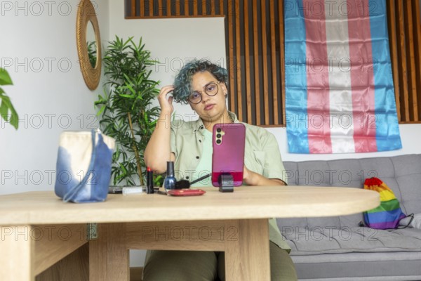 A transgender man sits at a wooden table, engaging in a makeup tutorial using his mobile phone. With pride flag in the background, he is surrounded by makeup products