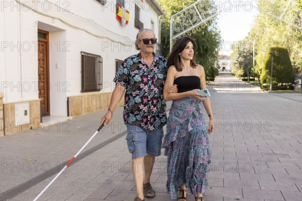 A blind father, utilizing a white cane, walks confidently through a town, guided by his attentive daughter on a sunny day