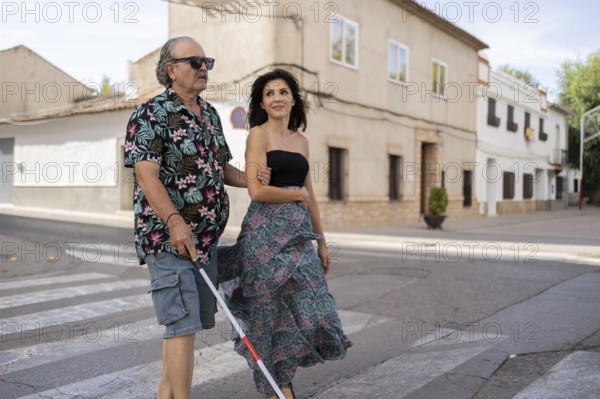 A blind father uses a white cane while his daughter guides him through a tranquil town street. Both dressed in casual summer attire, they navigate the surroundings with a sense of confidence and ease