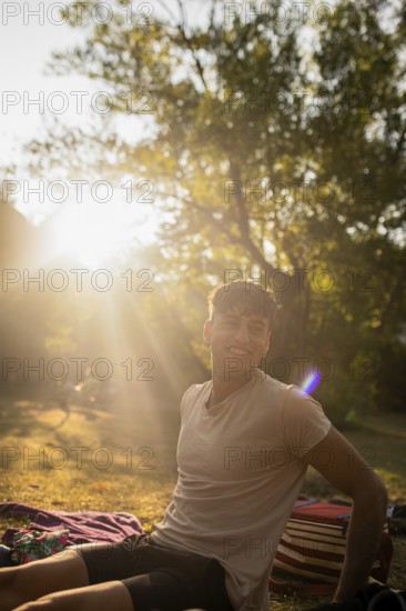 A young man relaxes outdoors by a lake, smiling broadly as he enjoys the warm summer sunshine filtering through trees, creating a cheerful, idyllic scene during sunset