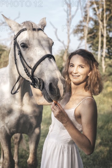 Young woman in a white dress gently pets a white horse on a sunny day. The serene outdoor setting captures a moment of friendship and tranquility in nature