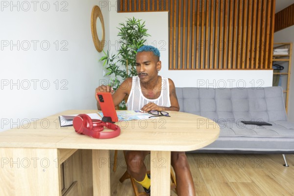 A Hispanic LGBTQIA+ man with vibrant style sits at a table, utilizing a smartphone and maps for holiday planning. Their focused expression shows anticipation