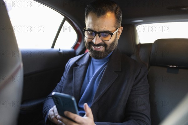 A happy Indian man, looking down at his smartphone, sits in a car, dressed in a winter coat and eyeglasses