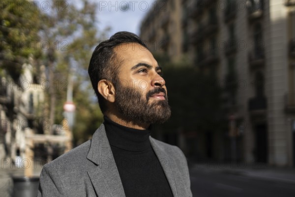 A confident Indian man dressed in a business-appropriate gray coat and black turtleneck is captured gazing upwards on a sunlit city street, embodying a classic winter style