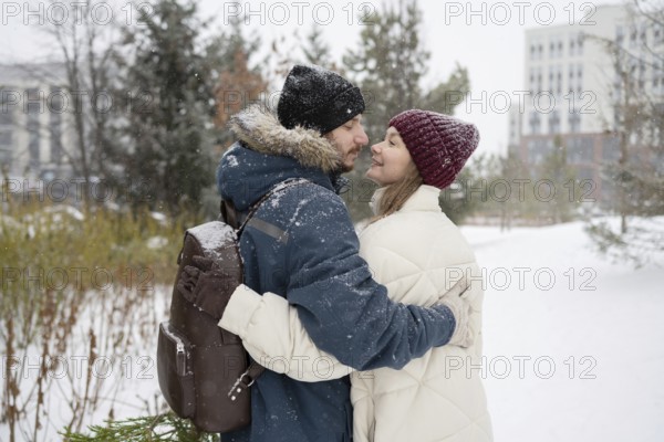 A couple warmly embraces in a snowy outdoor setting, surrounded by trees They wear winter attire with smiles and affection, creating a cozy and romantic atmosphere