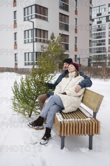 A couple sits on a wooden bench surrounded by snow, holding a small Christmas fir They are bundled up in winter clothing, enjoying the serene winter setting