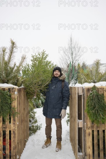 A man dressed warmly in a blue coat and gloves holds a fresh Christmas fir Snowflakes fall gently, creating a serene winter holiday atmosphere
