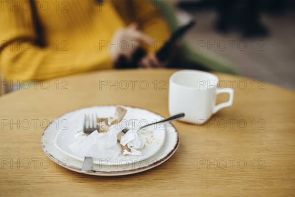 A cozy coffee shop setting featuring a wooden table with a used cup and plate Crumpled paper napkins add a lived-in touch A person in a yellow sweater is visible in the background, holding a phone