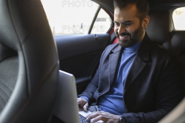 A happy Indian man dressed in professional attire works on his laptop while sitting in the backseat of a car, showcasing a modern, mobile business lifestyle