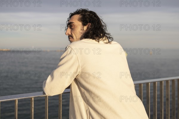 A man with long hair stands by a railing, gazing at the serene ocean under soft sunlight, conveying a sense of calm and introspection The horizon stretches in the distance