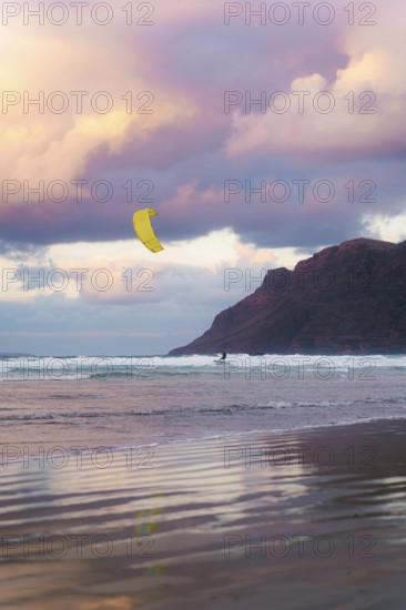 A kite surfer glides across the waves at Famara Beach, Lanzarote, under a sky painted with stunning sunset hues. The dramatic cliffs add to the breathtaking scene
