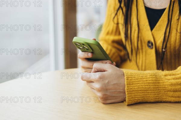 A person in a cozy yellow sweater holds a green smartphone while sitting at a wooden table near a window The focus is on the phone, highlighting modern technology