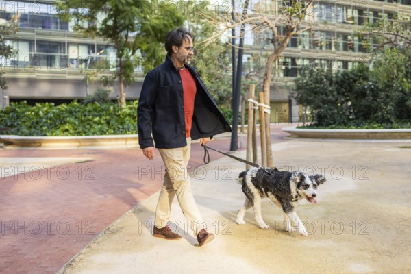 A mature man strolls with a dog through an urban park, both enjoying a sunny day. They are surrounded by modern architecture and greenery, depicting a peaceful everyday city life scene