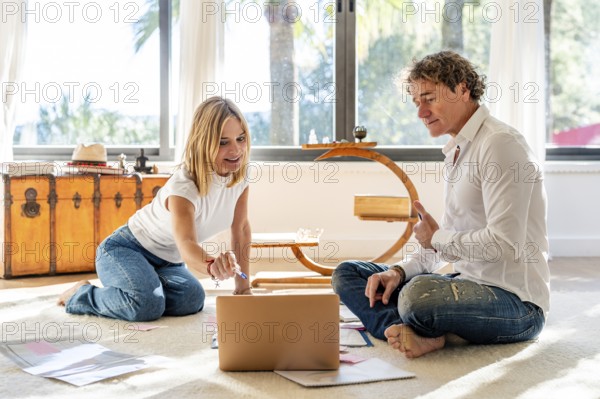 A man and woman sitting on the floor at home, engaging in work and planning activities. The man and woman are looking at a laptop and papers while discussing, in a bright, sunlit room