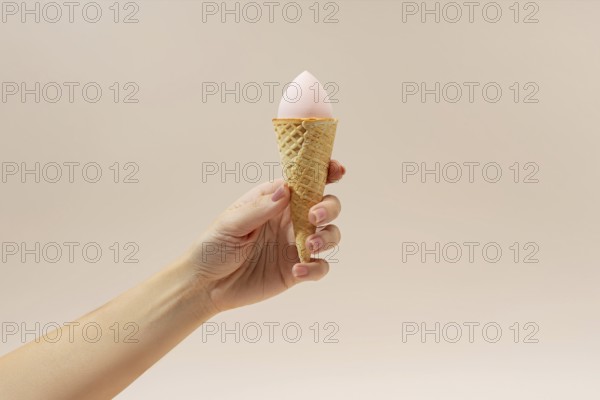 A hand holds a waffle cone with a pink cosmetic sponge inside, resembling ice cream The neutral background enhances the creative contrast of this beauty-themed image