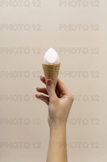 A hand holding a waffle cone with a cosmetic sponge inside, set against a soft, neutral background The image creatively blends beauty and everyday items