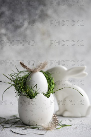 A meticulously crafted Easter setting featuring a white speckled egg cradling a nest of fresh green grass, topped with a delicate feather, with a sleek white ceramic bunny nearby, set against a soft, textured gray background