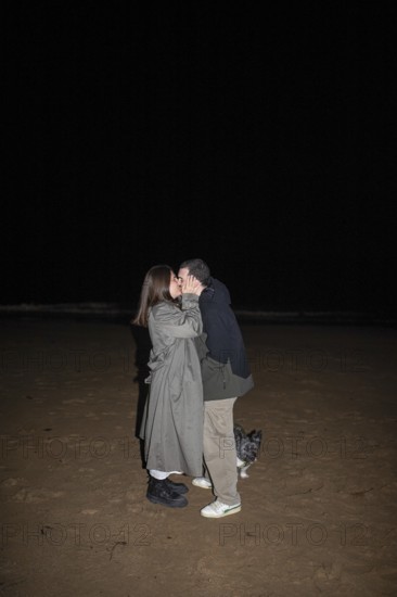 A couple shares a tender kiss under the night sky on a sandy beach, accompanied by a small dog The intimate moment captures love, tranquility, and the essence of companionship