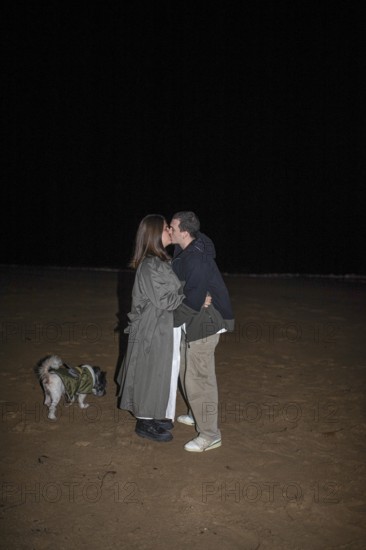 A couple shares a tender kiss on a dark beach night, their small dog beside them The scene captures romance and tranquility, with the vast night sky as a backdrop