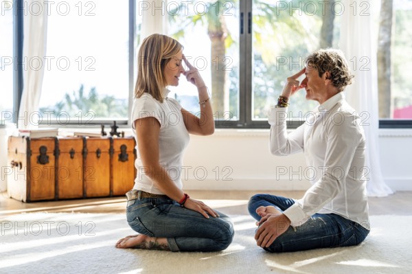 A man and woman meditate in a brightly lit room, utilizing alternative work strategies. The woman closes her eyes with fingers on temples while the man mirrors the pose, both in casual attire