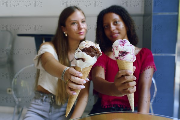 Two teenage friends, a Caucasian girl and an African girl, happily holding and showcasing their ice cream cones