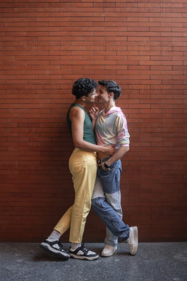 A young gay couple shares an intimate moment against a brick wall, expressing affection and love. Their casual attire and joyful expressions convey a sense of happiness